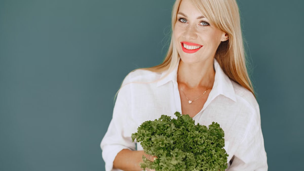 Cheerful woman with blonde hair holding fresh lettuce, promoting healthy living in a studio setting.